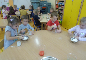 Czwórka dzieci siedzi przy stole, jedna dziewczynka nabiera łyżeczką sól z miski, reszta dzieci rozpuszcza sól w wodzie.